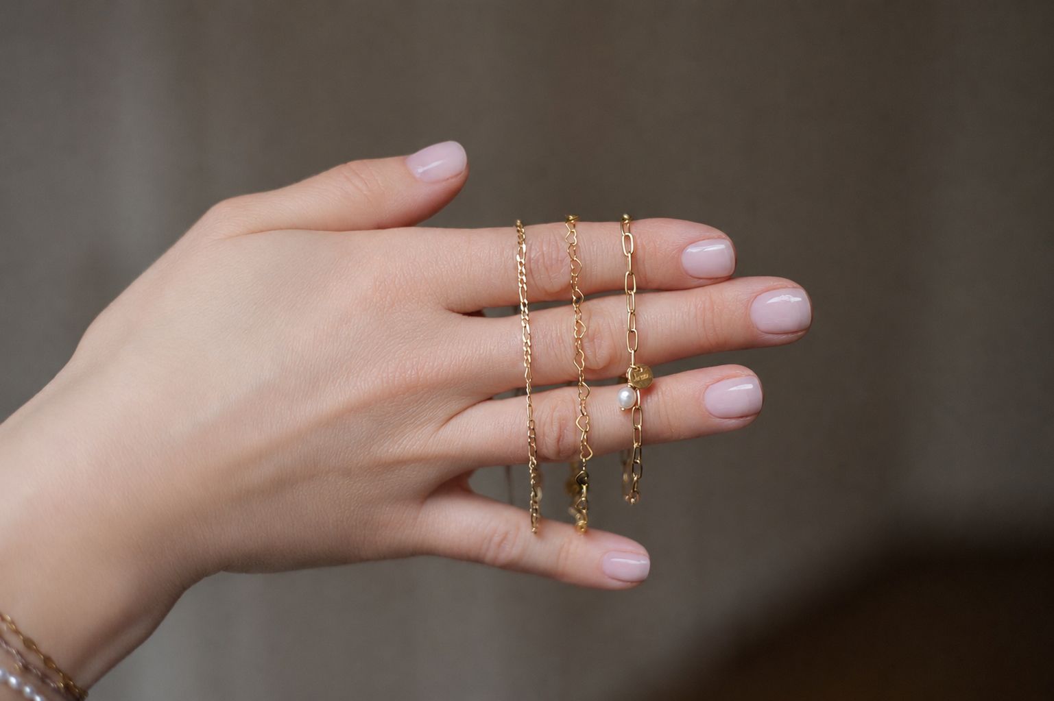 Drei goldene Armbänder über einer Hand — Figarokette, Herzchenkette und Gliederkette mit Perle